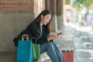 Güzel bir Asyalı kadın bir alışveriş merkezinde şık bir şekilde dolaşırken, alışveriş torbaları taşırken ve şık bir kıyafet giyerken akıllı telefonunun keyfini çıkarırken görülüyor..