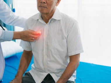 Asyalı bir erkek doktor steteskoplu yaşlı bir hastayı muayene ediyor, rutin bir sağlık kontrolü yapıyor. Erkek hasta klinikte gözlem için uzanıyor..