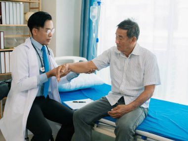 Asyalı bir erkek doktor ve yaşlı bir hasta, hastane koğuşunda hastaya yardım etmek için fizyoterapi yapmak üzere el ele tutuşur..