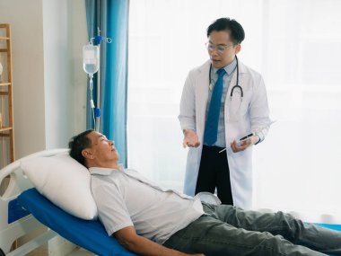 Asyalı bir erkek doktor, hasta yatağında yatan yaşlı bir erkek hastayla konuşuyor. Hastanın odasında sağlık muayeneleri ve sağlık kontrolleri yapılıyor..