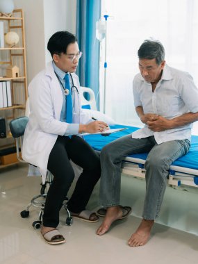 Asyalı bir erkek doktor ve yaşlı bir erkek hasta, bir sağlık konsepti kliniğinde yatakta otururken zehirlenmenin yol açtığı mide ağrısını tartışıyorlar..