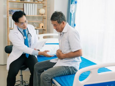 Asyalı bir erkek doktor ve yaşlı bir erkek hasta, bir sağlık konsepti kliniğinde yatakta otururken zehirlenmenin yol açtığı mide ağrısını tartışıyorlar..