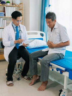 Asyalı bir erkek doktor ve yaşlı bir erkek hasta, bir sağlık konsepti kliniğinde yatakta otururken zehirlenmenin yol açtığı mide ağrısını tartışıyorlar..