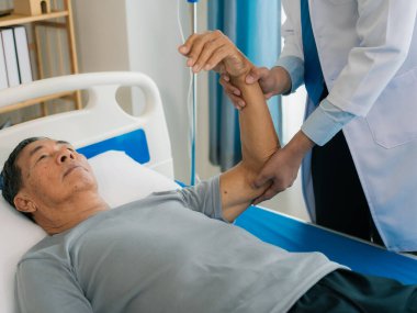 Asyalı bir erkek doktor ve yaşlı bir hasta, hastane koğuşunda hastaya yardım etmek için fizyoterapi yapmak üzere el ele tutuşur..