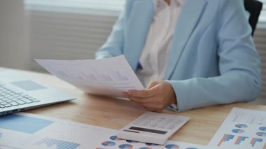 Asyalı bir iş kadını masasındaki mali raporları analiz ediyor. Harcamaları, grafikleri ve dizüstü bilgisayarını kullanarak gelir ve giderleri hesaplıyor..