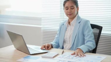 Asyalı bir iş kadını masasındaki mali raporları analiz ediyor. Harcamaları, grafikleri ve dizüstü bilgisayarını kullanarak gelir ve giderleri hesaplıyor..
