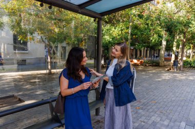 Otobüs durağındaki gülümseyen kadınlar birlikte şehir haritası okurlar, gezi yolculuklarının tadını çıkarırlar..