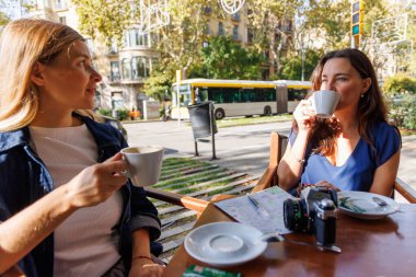 Kafe terasında konuşan ve gülen iki kadının doğal anı, şehirde bağlantı ve neşe gösteriyor..