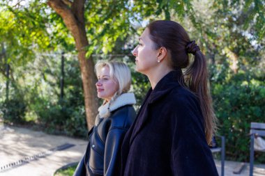 İki arkadaş açık bir parkta yavaşça yürüyor, rahatça konuşuyorlar ve dışarıda huzurlu bir anın tadını çıkarıyorlar..