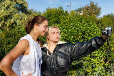 İki arkadaş, parlak ve sakin bir parkta yürürken eski bir kamerayla eğlenceli selfie çekiyorlar..