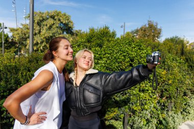 İki kadın, doğal ve huzurlu bir park ortamında yürürken klasik bir kamerayla gülümsüyor ve selfie çekiyor..