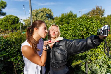 İki kadın, güneşli ve yeşil bir parkta rahat bir yürüyüş yaparken yaratıcı selfie 'ler için klasik bir kamera kullanıyor..