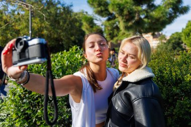 İki kadın, eğlence amaçlı selfie çekmek için eski bir kamerayı sırayla kullanıyor. Parkta rahat ve dostça bir yürüyüşün keyfini çıkarıyorlar..