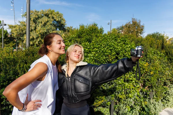 İki kadın, doğal ve huzurlu bir park ortamında yürürken klasik bir kamerayla gülümsüyor ve selfie çekiyor..