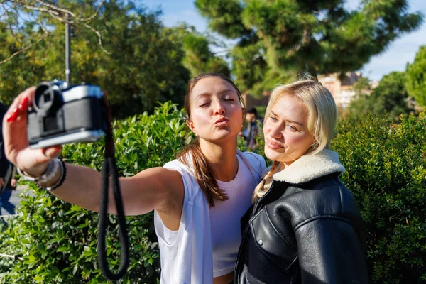 İki kadın, eğlence amaçlı selfie çekmek için eski bir kamerayı sırayla kullanıyor. Parkta rahat ve dostça bir yürüyüşün keyfini çıkarıyorlar..