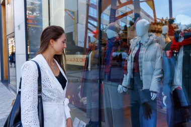 Yumuşak alışveriş merkezinin ışıklarının altında duruyor, elinde birkaç renkli çanta, üst düzey vitrinlere dik dik bakıyor.