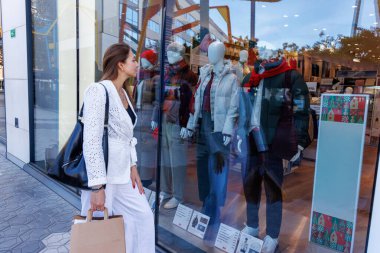 Modayı takip eden bir alışveriş merkezinin içinde, elinde çantalar, tuhaf bir şekilde şık eşyalarla dolu butik pencerelere bakıyor.