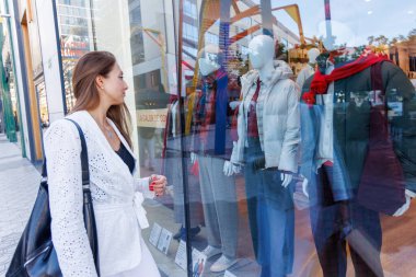 Alışveriş torbalarıyla dolu alışveriş merkezleri koridorlarında dolaşır, moda ve yaşam tarzı ürünleriyle dolu pencerelere bakar.