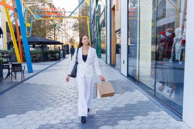 Parlak bir alışveriş merkezinin içinde elinde renkli çantalarla geziyor, güncel moda eğilimleriyle dolu vitrinleri değerlendiriyor.