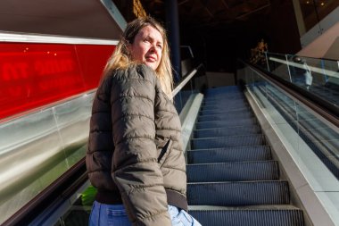 Genç, çekici, sarışın bir kadın ticari bir binada yürüyen merdiven kullanıyor. Moda, ulaşım ve kentsel eğlence için ideal yüksek kaliteli fotoğrafçılık.