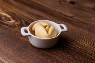 Kırsal bir ahşap yüzeyde seramik beyaz bir kasede otantik şans kurabiyeleri. Dramatik ışıklandırma, şans ve refahın simgelerini vurguluyor..