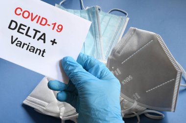 Doktorun elinde beyaz kağıtlı mavi eldiven ve Covid-19 Delta + Variant 'a arka planda çeşitli koruma maskeleri olan mesaj var. COVID-19 delta değişkeni tür koruma kavramı.