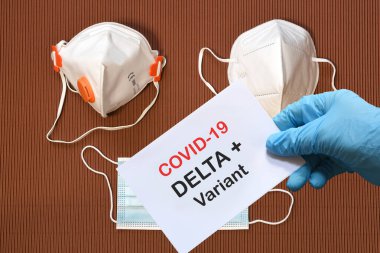 Doktorun elinde beyaz kağıtlı mavi eldiven ve Covid-19 Delta + Variant 'a arka planda çeşitli koruma maskeleri olan mesaj var. COVID-19 delta değişkeni tür koruma kavramı.