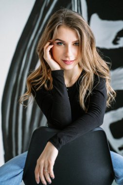 Siyah tişörtlü ve kot pantolonlu, sarı saçlı güzel bir kızın portresi bir duvarın arkasında oturan zebra desenli siyah bir sandalyede.