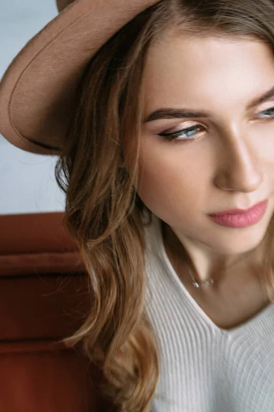 Una bella ragazza bionda con un cappello si siede su un divano marrone in un elegante appartamento bianco — Foto Stock