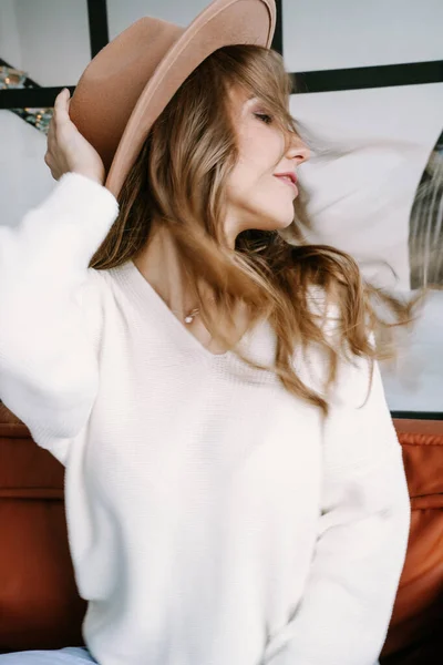 Una hermosa chica rubia en un sombrero se sienta en un sofá marrón en un elegante apartamento blanco — Foto de Stock