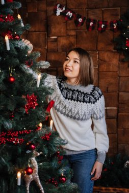 Kısa saçlı esmer bir kız, gri kazaklı bir yılbaşı ağacının yanında oturuyor..