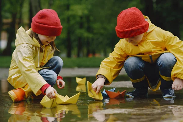 Funny kids in rain boots playing with paper ship by a puddle — Stock Photo, Image