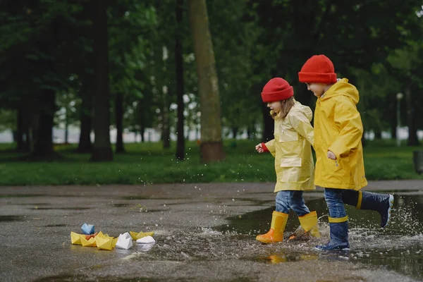 Funny kids in rain boots playing with paper ship by a puddle — Stock Photo, Image