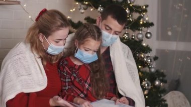 Mutlu beyaz maskeli aile, Noel arifesinde Noel ağacının yanındaki süslü evde birlikte oturuyorlar.
