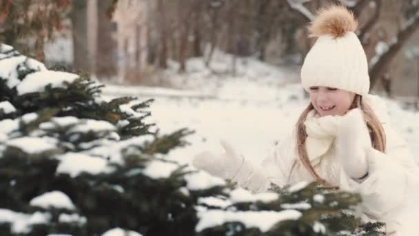 Skolflicka i vita vinterkläder står nära granen täckt med snö — Stockvideo