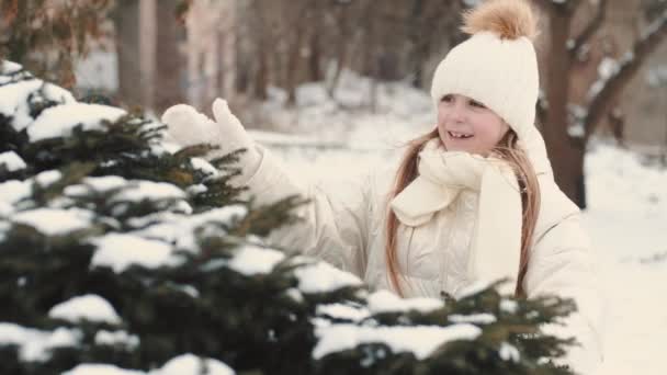 Skolflicka i vita vinterkläder står nära granen täckt med snö — Stockvideo