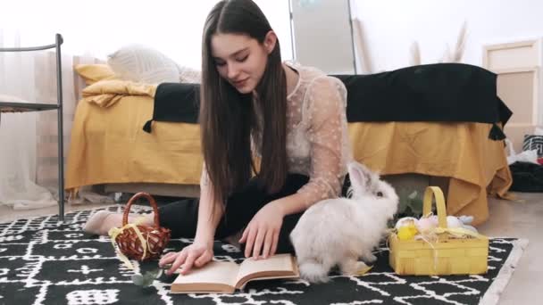 Дівчина сидить читаючи книгу з кроликом на підлозі — стокове відео