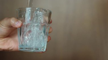 İçinden su akarken temiz bir bardağı tutan bir el hareketi ve berraklığı yakalıyor. Arka plan yumuşakça bulanık, hidrasyona ve şeffaf sıçramaya odaklanmış.