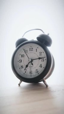 Ahşap yüzey ve parlak beyaz arka plan üzerinde klasik bir siyah alarm saatinin dikey görüntüsü. Zaman yönetimi kavramları, çalışma saatleri ve zaman hızla geçiyor.