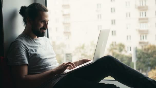 Joven hombre guapo trabajando en su portátil mientras está sentado en un alféizar de la ventana — Vídeos de Stock