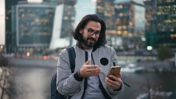 Beau jeune homme barbu dans des lunettes de prendre un appel vidéo avec son smartphone — Video