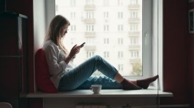 Genç bir kadın pencere kenarında oturuyor, cep telefonu kullanıyor ve kahve içiyor.