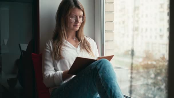 Uma jovem senta-se no peitoril da janela, lê um livro e bebe café de uma caneca — Vídeo de Stock