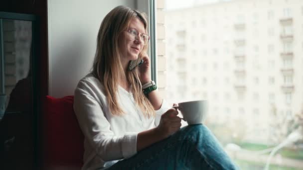 En ung kvinna sitter på fönsterbrädan och pratar i mobilen. — Stockvideo