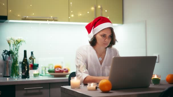 Uma jovem mulher de chapéu de Pai Natal está digitando em um laptop e bebendo champanhe — Vídeo de Stock