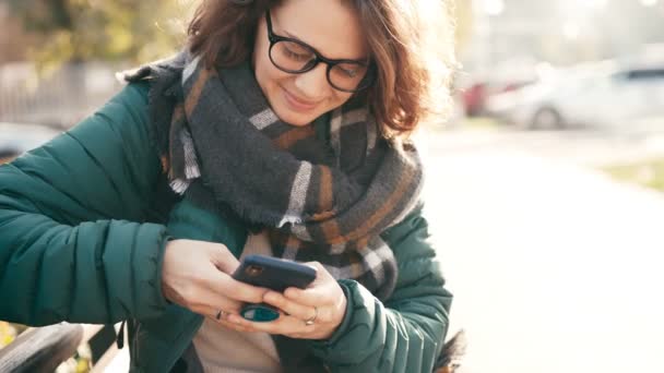 Молодая веселая женщина в очках сидит на скамейке со смартфоном в руках — стоковое видео