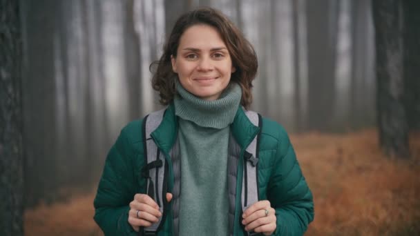 Молодая веселая женщина стоит на фоне леса и смотрит в камеру — стоковое видео