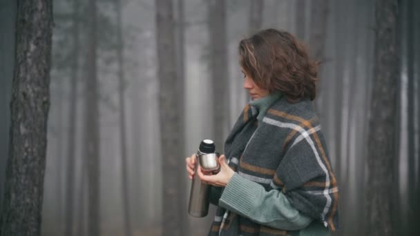 大きな居心地の良いスカーフに包まれた若い女性は魔法瓶からお茶を飲みます — ストック動画