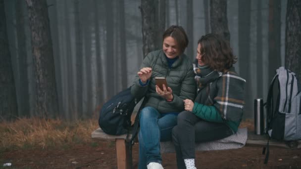 Két fiatal, vidám nő szelfit készít egy okostelefonon egy őszi erdőben. — Stock videók