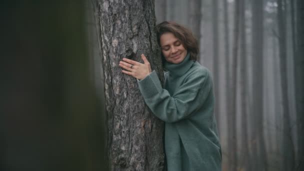 En ung kvinna i en varm, mysig tröja lutad mot trädstammen i en skog — Stockvideo